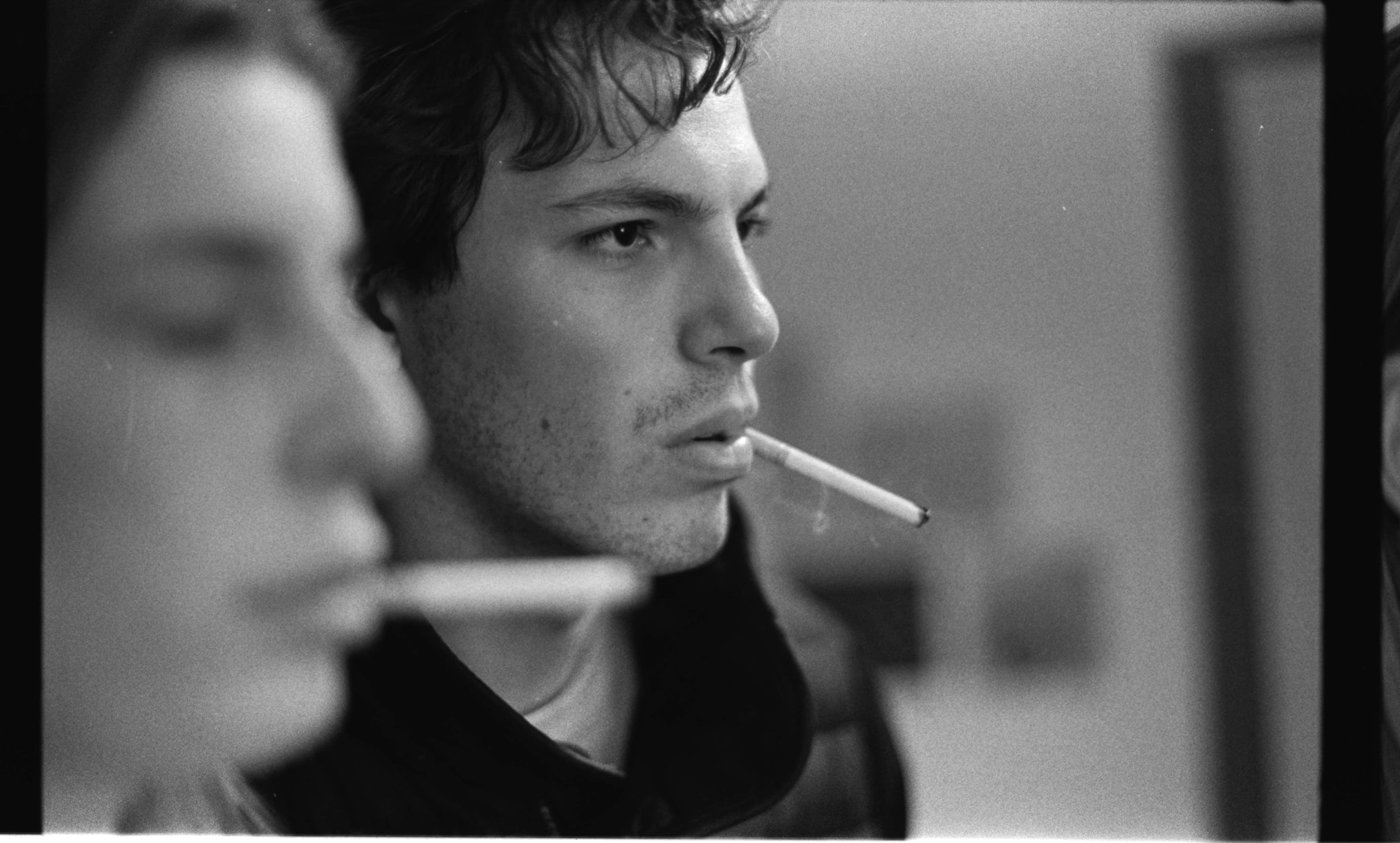 Person smoking a cigarette, looking reflective, with another person’s face partially seen in the foreground.