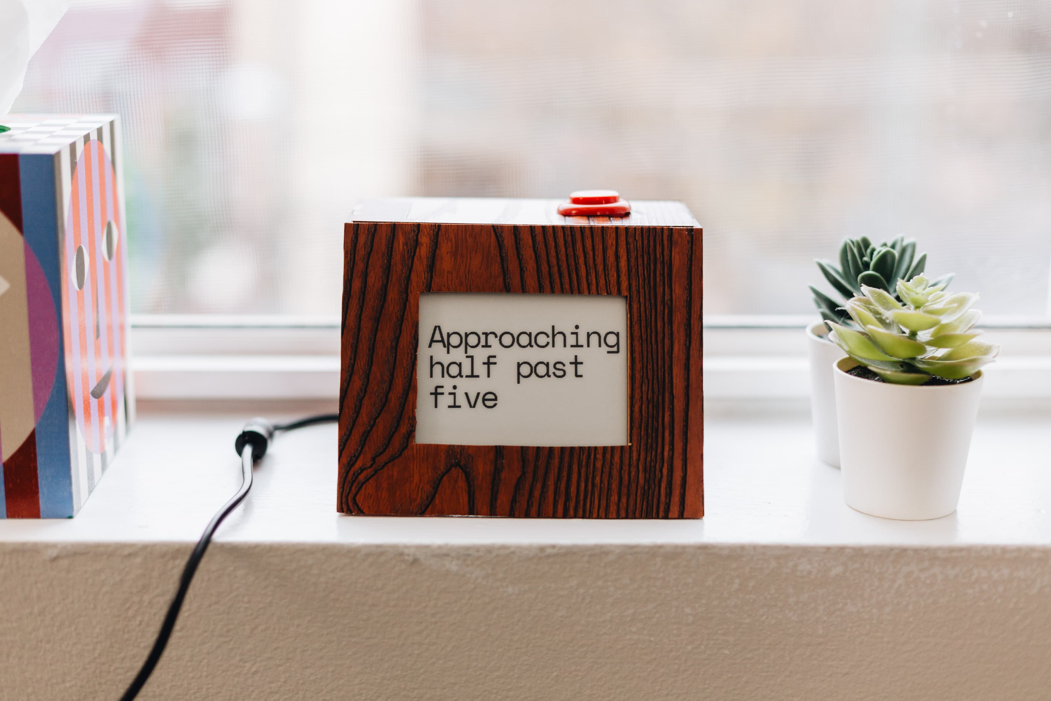 The fuzzy clock in a dark wood-grain enclosure on a windowsill, displaying "Approaching half past five" on its e-paper screen, with a red button on top and a small succulent beside it