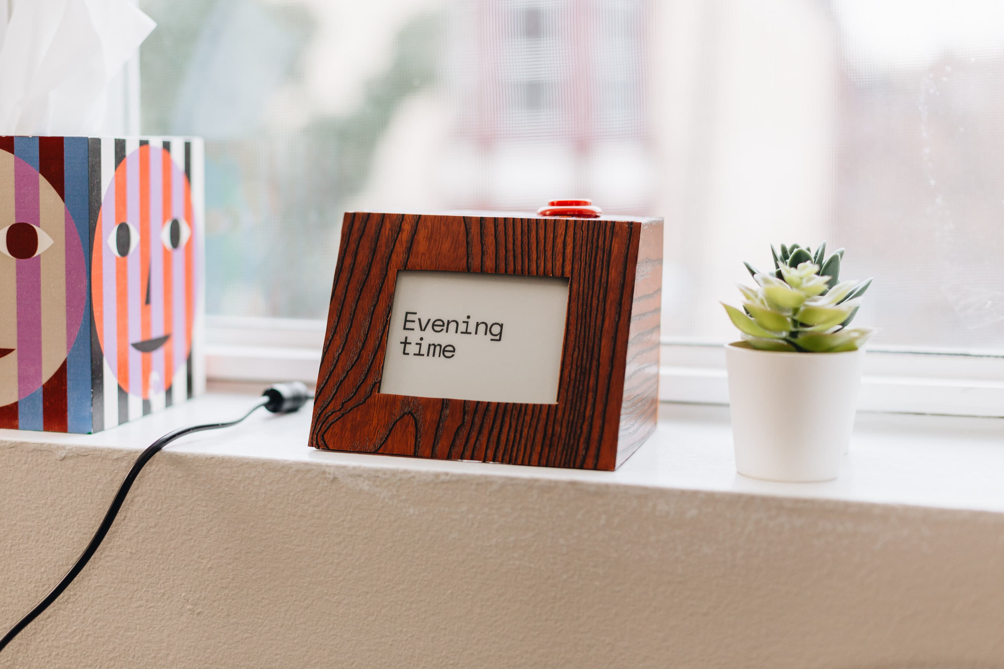 The fuzzy clock in its dark wood-grain enclosure on a windowsill, displaying "Evening time" on its e-paper screen, next to a small succulent and colorful art prints